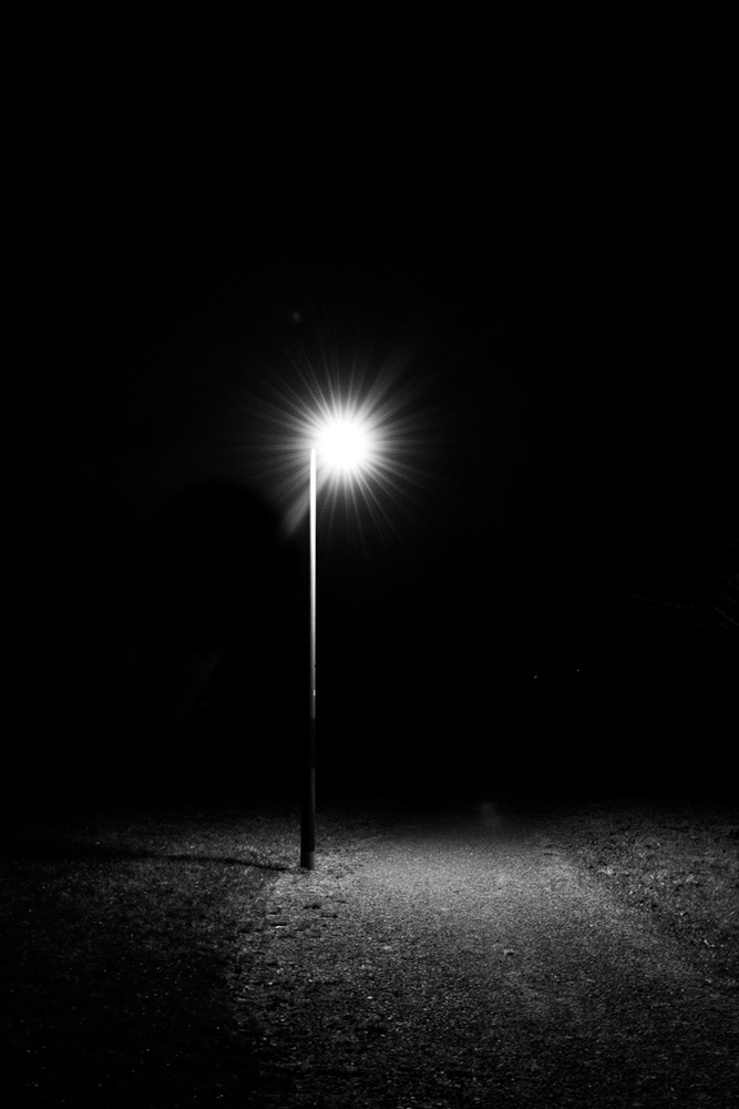 Schwarzweiß Foto ein Laterne, dahinter ein dunkler Weg.